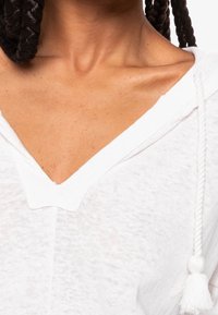Biały, lekki hoodie z dekoltem w serek, teksturowanym materiałem i sznurkiem z frędzlami. Zbliżenie na dekolt i okolicę ramion.