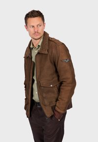 Giacca in pelle marrone con chiusura a zip, due tasche frontali e polsini a costine. Presenta una toppa sulla spalla e un colletto strutturato.