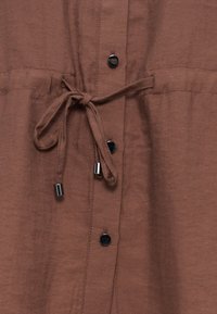 Tissu marron avec boutons-pression noirs et cordon noué avec embouts métalliques argentés au centre.