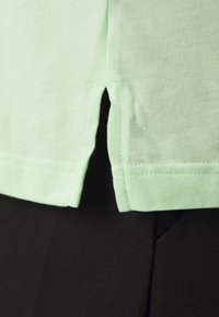 Lys grøn bomuld t-shirt med en lille sideslids og syet kant, parret med sort stof nedenunder. Glat tekstur, enkelt design.