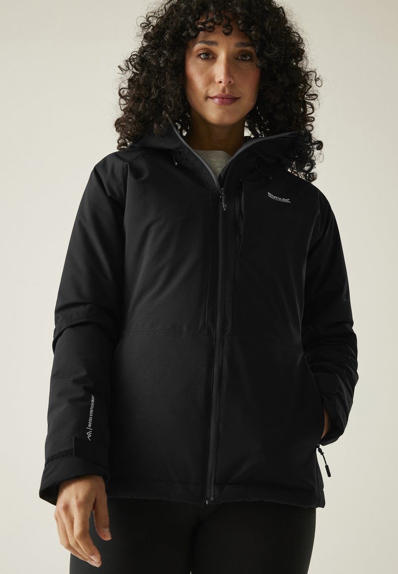 Femme aux cheveux bouclés portant une veste d'extérieur noire à capuche avec fermeture éclair et poches à main, debout devant un fond clair.