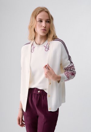 Giovane donna che indossa un cardigan bianco con bottoni sopra un maglione bianco con ricamo floreale viola e pantaloni viola, in piedi davanti a uno sfondo semplice.