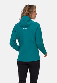 Giacca soft-shell verde acqua con cappuccio, dotata di una ventilazione posteriore, maniche lunghe e un logo discreto. Indossata con pantaloni neri attillati.