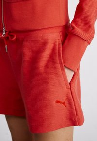 Shorts rouges en tissu texturé avec une taille à cordon, des poches latérales et un petit logo Puma orange en bas à droite.