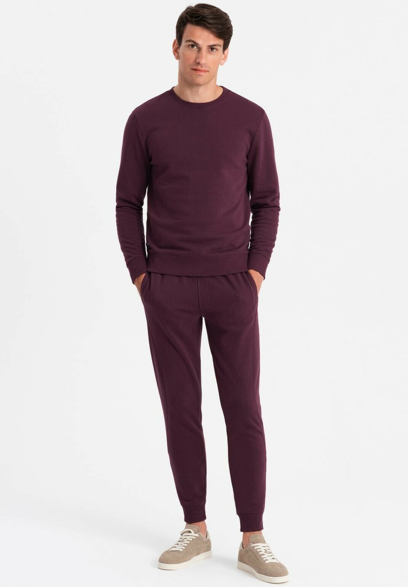 Lila Baumwoll-Sweatshirt mit gerippten Bündchen und passenden Jogginghosen, die einen lockeren Schnitt und eine glatte Textur aufweisen. Beigefarbene Sneakers runden den Look ab.