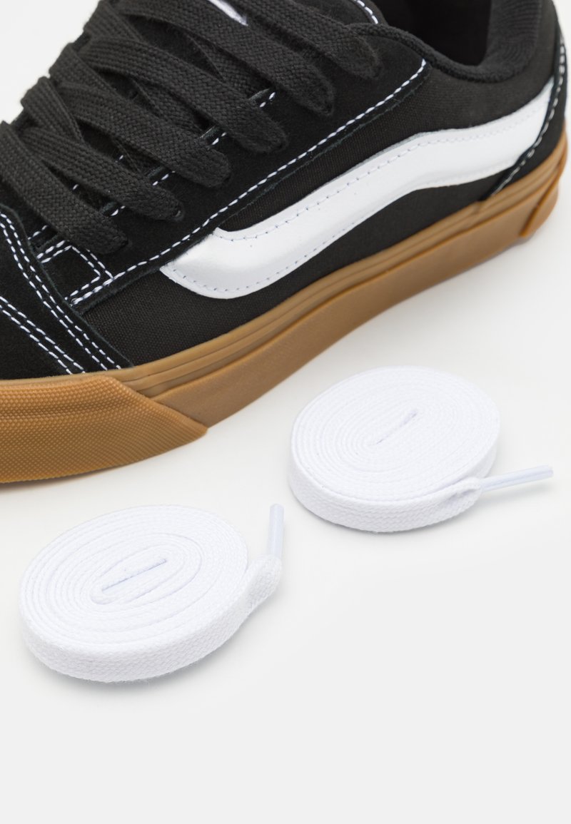 Sneaker nera in tela con striscia laterale bianca e suola in gomma color caramello. Due lacci bianchi a spirale sono posizionati nelle vicinanze.