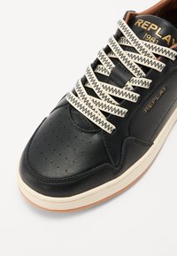 Sneaker din piele neagră cu șireturi crem contrastante, zonă perforată la vârf, talpă din cauciuc gumă și branding subtil aurit pe lateral.