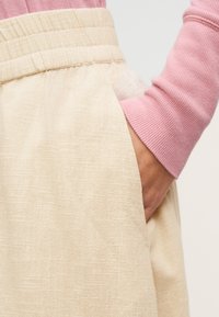 Χέρι στην τσέπη μπεζ υφασμάτινου παντελονιού με ελαστική μέση, φορώντας ροζ μακρυμάνικη ριμπ μπλούζα σε απλό φόντο.