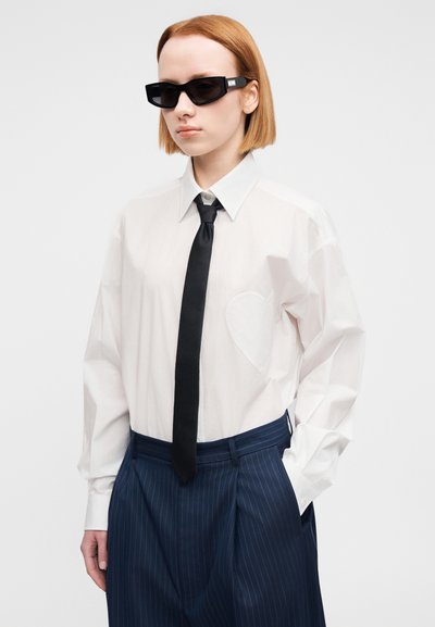 Λευκό πουκάμισο με κουμπιά και τσέπη σε σχήμα καρδιάς, μαύρη γραβάτα και σκούρο παντελόνι με λεπτές ρίγες. Το μοντέλο φοράει μαύρα γυαλιά ηλίου.