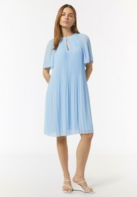 Helles blaues, plissiertes Kleid mit Rundhalsausschnitt und Schlüsselloch-Ausschnitt. Verfügt über fließende Ärmel und eine schicke Textur, kombiniert mit weißen Sandalen mit Riemen.