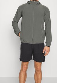 Chaqueta ligera con cremallera en tela gris oscuro, con capucha, mangas largas y diseño ajustado. Combinada con pantalones cortos negros con franjas laterales.