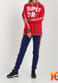 Rode hoodie met grote witte tekst "SUPER DR", marineblauwe joggingsbroek met witte zijkant en zwarte sportieve schoenen. Casual sportieve outfit.