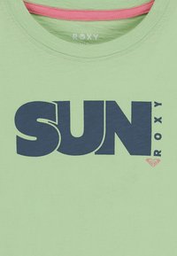 Světle zelené tričko s kulatým výstřihem, které má výrazný námořnický text "SUN" a menší logo "ROXY" v růžové barvě v dolním rohu.