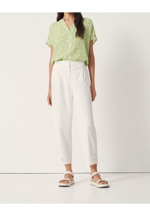 Kobieta w jasnooliwkowej bluzce w krótkie rękawy z wzorem, białych spodniach z wysokim stanem i paskami przy kostkach oraz beżowych sandałach stoi na białej podłodze.