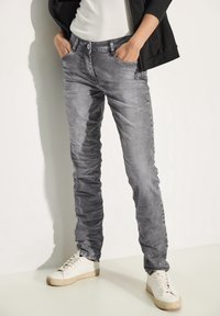 Jean en denim gris avec une coupe slim, présentant des poches avant, une texture subtile, et associés à des baskets blanches sur un fond uni.