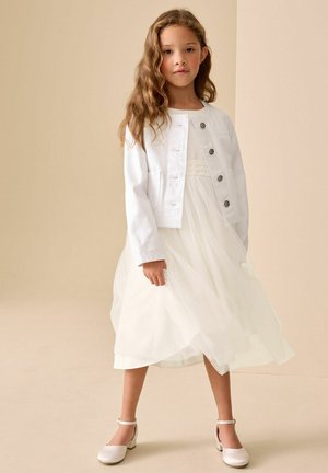 Pikkade laineliste juustega noor tüdruk, kes kannab valget tuletuskangast kleiti, valget nööbitud jopet ja valgeid pahkluu rihmaga kingi, seisab beežil taustal.