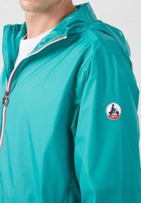 Turquoise waterdichte jas met capuchon, ritssluiting aan de voorkant en elastische manchetten. Voorzien van een logopatch op de linkerarm. Gladde textuur.