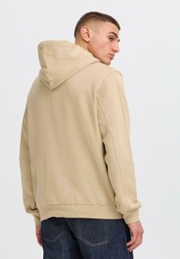 A young man with short hair wears a beige hoodie, looking over his shoulder. The background is plain and light-colored.
