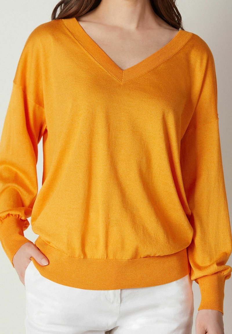 Orange V-halsad pullover i mjukt tyg med långa ballongärmar och ribbat, åtsittande avslut. Bärs över vita byxor och visar en avslappnad design.