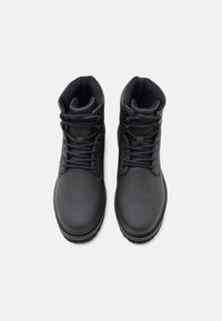 Un paio di stivali neri alla caviglia con lacci e colletto imbottito, visti dall'alto su uno sfondo chiaro e uniforme.