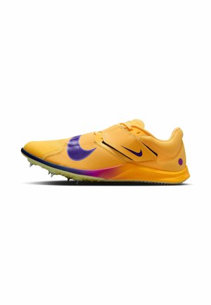 Chaussure de sport jaune avec un swoosh Nike violet, dessus texturé, fermeture par sangle et semelle en caoutchouc à crampons pour une meilleure adhérence.