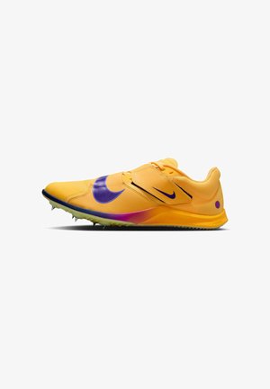 Chaussure de sport jaune avec un swoosh Nike violet, dessus texturé, fermeture par sangle et semelle en caoutchouc à crampons pour une meilleure adhérence.