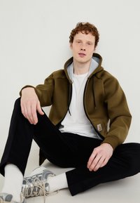 Olivnozeleni zip-up kapucar z sivim notranjim delom kapuce, črnimi vrvicami in logotipom; nošen preko belega majic in kombiniran s črnimi hlačami.