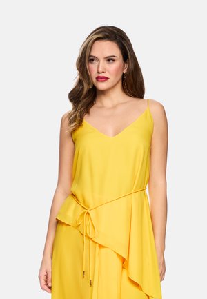 Woman with long wavy hair wears sleeveless yellow dress with thin straps and waist tie, looking slightly to the side against a plain background.