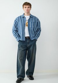 Camicia a quadri blu con bottoni sopra una maglietta bianca con grafica, abbinata a jeans scuri larghi e scarpe nere. Accessori semplici.