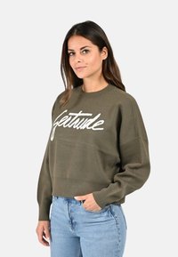 Pull en vert olive avec un design cropped, présentant le texte brodé blanc "Gesture" sur le devant. Tissu doux et coupe décontractée.