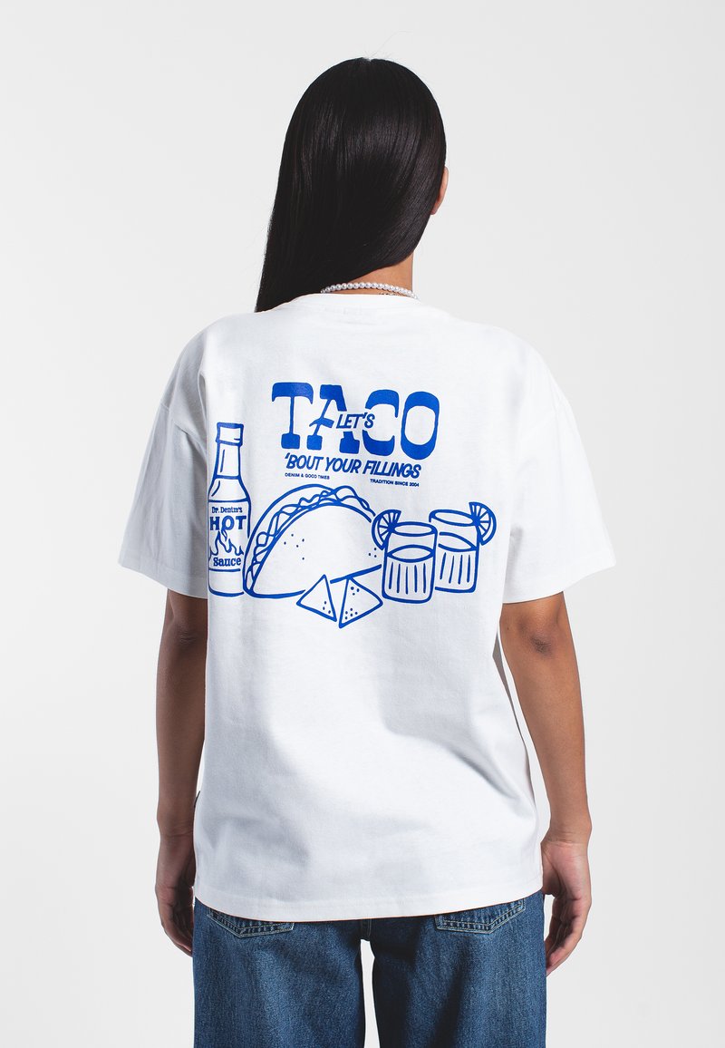 Bílý bavlněný tričko s modrým grafickým designem na zádech, které zobrazuje tacos, nápoje a láhev s ostrou omáčkou s textem.