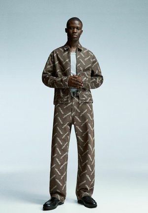 Young man standing with hands clasped wearing brown patterned jacket and pants set, white shirt, black belt, and black shoes on light background.