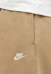 Κοντινό πλάνο μπεζ παντελονιών Nike με λευκή κεντημένη στάμπα κοντά στην πλαϊνή τσέπη κάτω από λευκό ένδυμα.