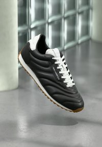 Black leather sneaker with white laces, textured side panels, and a gum rubber sole. Features a logo on the side, streamlined shape.