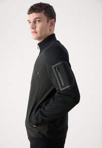 Black zip-up jacket featuring a smooth panel and textured sleeves, with side pockets and a zippered pocket on the left arm.