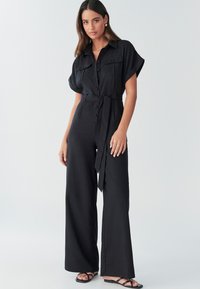 Mono negro con mangas cortas, cierre de botones en la parte delantera, dos bolsillos en el pecho, cinturón en la cintura, piernas amplias y combinado con sandalias negras. Textura suave.