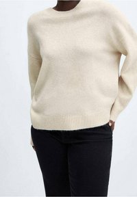 Pull en maille beige avec un col arrondi et un ourlet côtelé. Comprend des manches amples et une texture douce, associé à un jean noir.