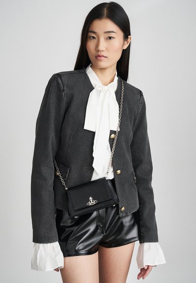 Casaco de ganga preto com botões dourados, usado sobre uma blusa branca com um laço, combinado com calções de couro preto brilhante e uma bolsa com textura de crocodilo.