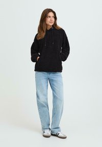 Sort fleece hættejakke med en tekstureret overflade, frontlomme og justerbare snore, parret med lyseblå jeans og hvide sneakers.