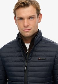 Navy puffer jacket with a zip closure, textured quilting, high collar, and a black logo patch featuring red accents on the chest.