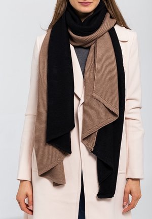 Femme portant un manteau beige et une longue écharpe tricotée avec des larges rayures verticales alternées noires et marron.