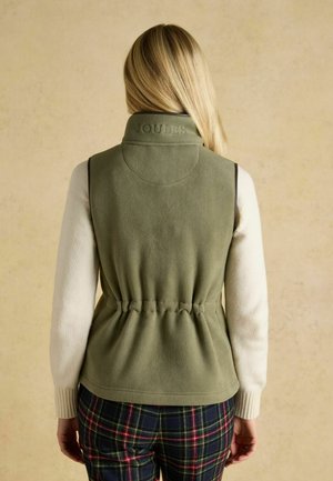 Blonde Frau trägt eine olivgrüne Fleeceweste mit "Joules" am Kragen, cremefarbenen Pullover und karierte Hose, blickt von der Kamera weg.