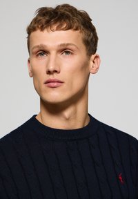 Marineblaue Kabelstrickpullover mit geripptem Rundhalsausschnitt, versehen mit einem kleinen roten Logoakzent. Weiche Textur, hervorgehobene Nähte.