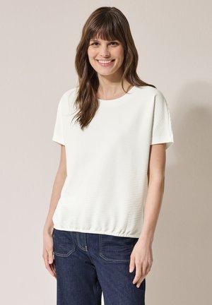 Top bianco a maniche corte con un motivo texturizzato e un collo rotondo. Abbinato a jeans in denim scuro con orlo arricciato.
