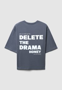 T-shirt grigio oversize con testo bianco in grassetto: "CANCELLA IL DRAMA TESORO" e testo più piccolo sopra e sotto, con scollatura rotonda.