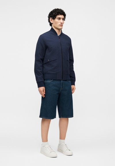 Homme portant une veste bomber bleu marine, un short en jean bleu au niveau des genoux, des chaussettes blanches et des baskets blanches, debout devant un fond uni.