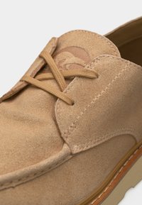 Κοντινή λήψη σε μπεζ σουέντ παπούτσι που δείχνει τα κορδόνια, τις λεπτομερείς ραφές και τη λαστιχένια σόλα με υφή για καλύτερο κράτημα.