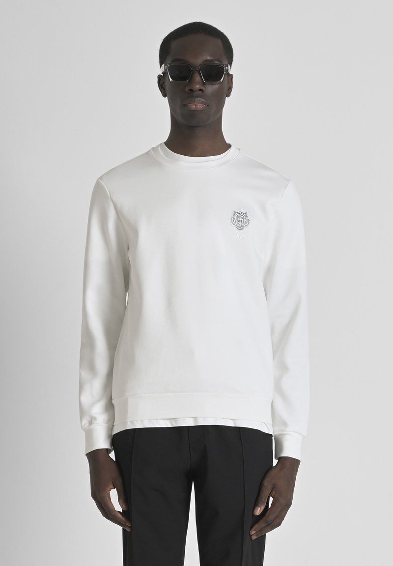 Sudadera blanca con un acabado texturizado, cuello redondo y logo bordado en el lado izquierdo del pecho. Combinada con pantalones negros.