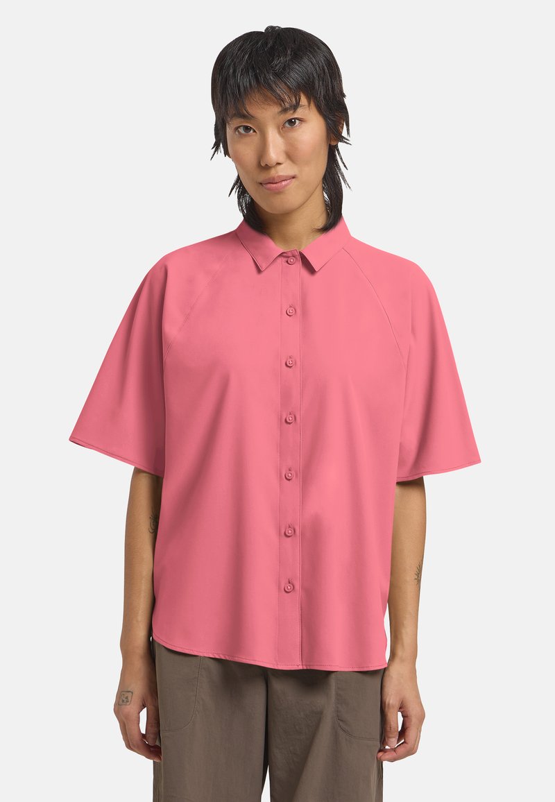 Personne aux cheveux courts et foncés portant une chemise rose ample boutonnée et un pantalon marron, sur un fond clair uni.