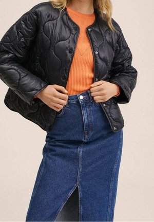 Femme portant une veste matelassée noire par-dessus un pull côtelé orange et une jupe en denim bleu taille haute avec une fente avant.